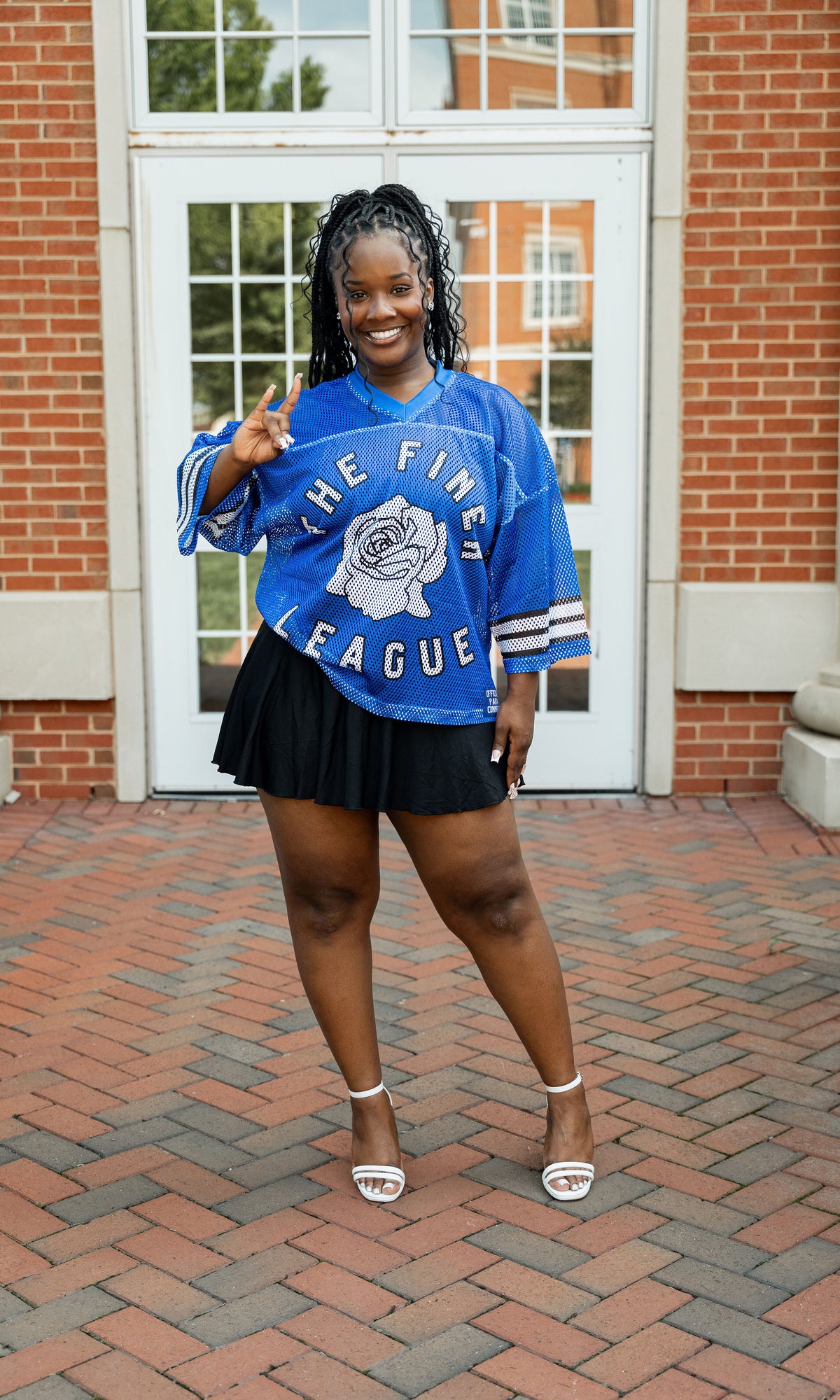 A woman wearing a blue mesh jersey with text 'THE FINEST LEAGUE' and stripes, paired with a black skirt and white shoes, standing on a brick pathway.