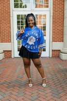 A woman wearing a blue mesh jersey with text 'THE FINEST LEAGUE' and stripes, paired with a black skirt and white shoes, standing on a brick pathway.