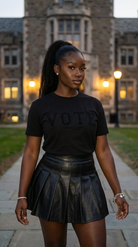Person wearing a black 'VOTE' t-shirt and leather skirt in front of a building.
